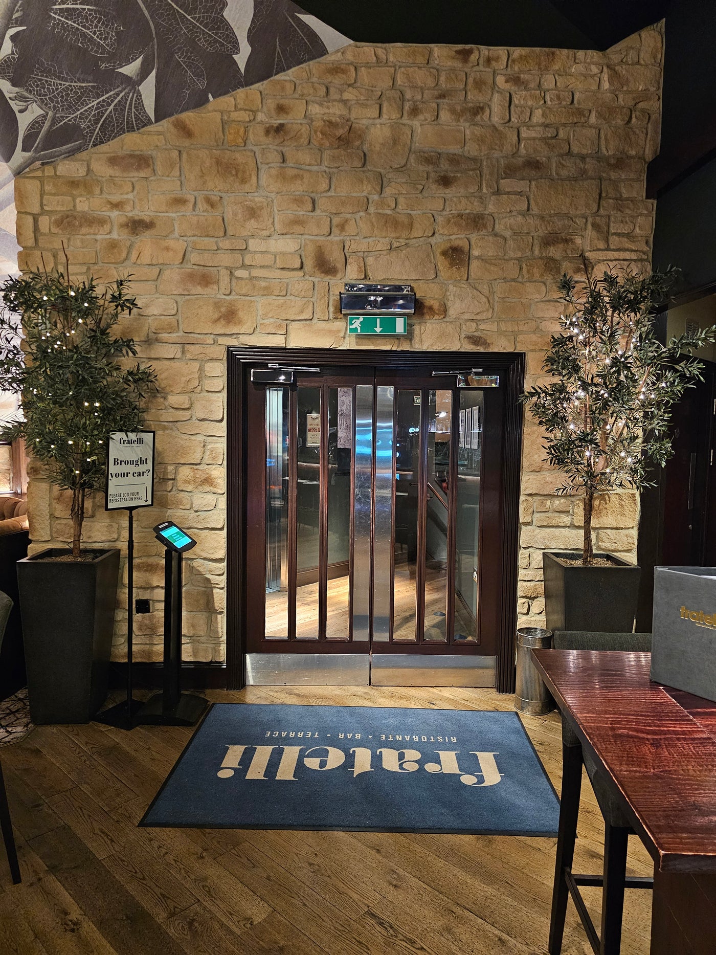 Entrance with stone cladding, wooden doors, and potted plants at a restaurant interior, featuring a welcome mat.