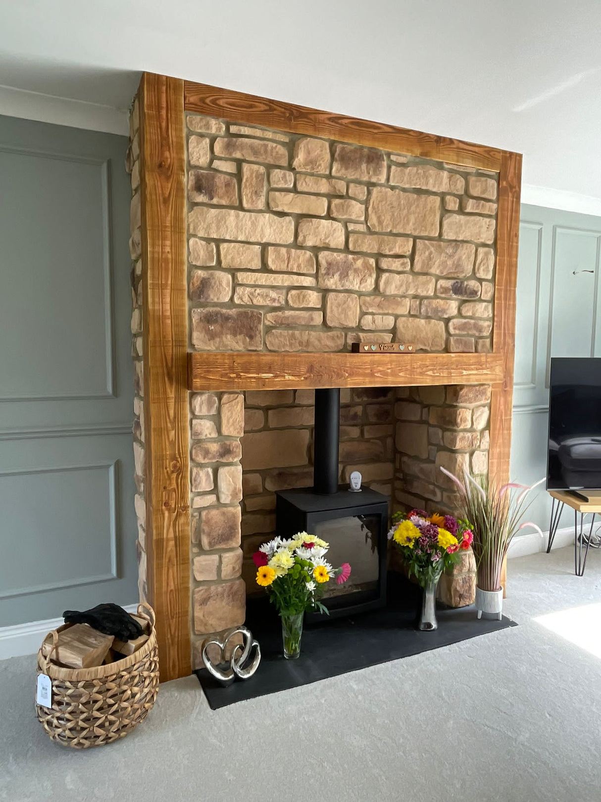 Stone cladding fireplace with wooden mantel and decorative flowers, showcasing Cotswold Blend design in a modern living room.