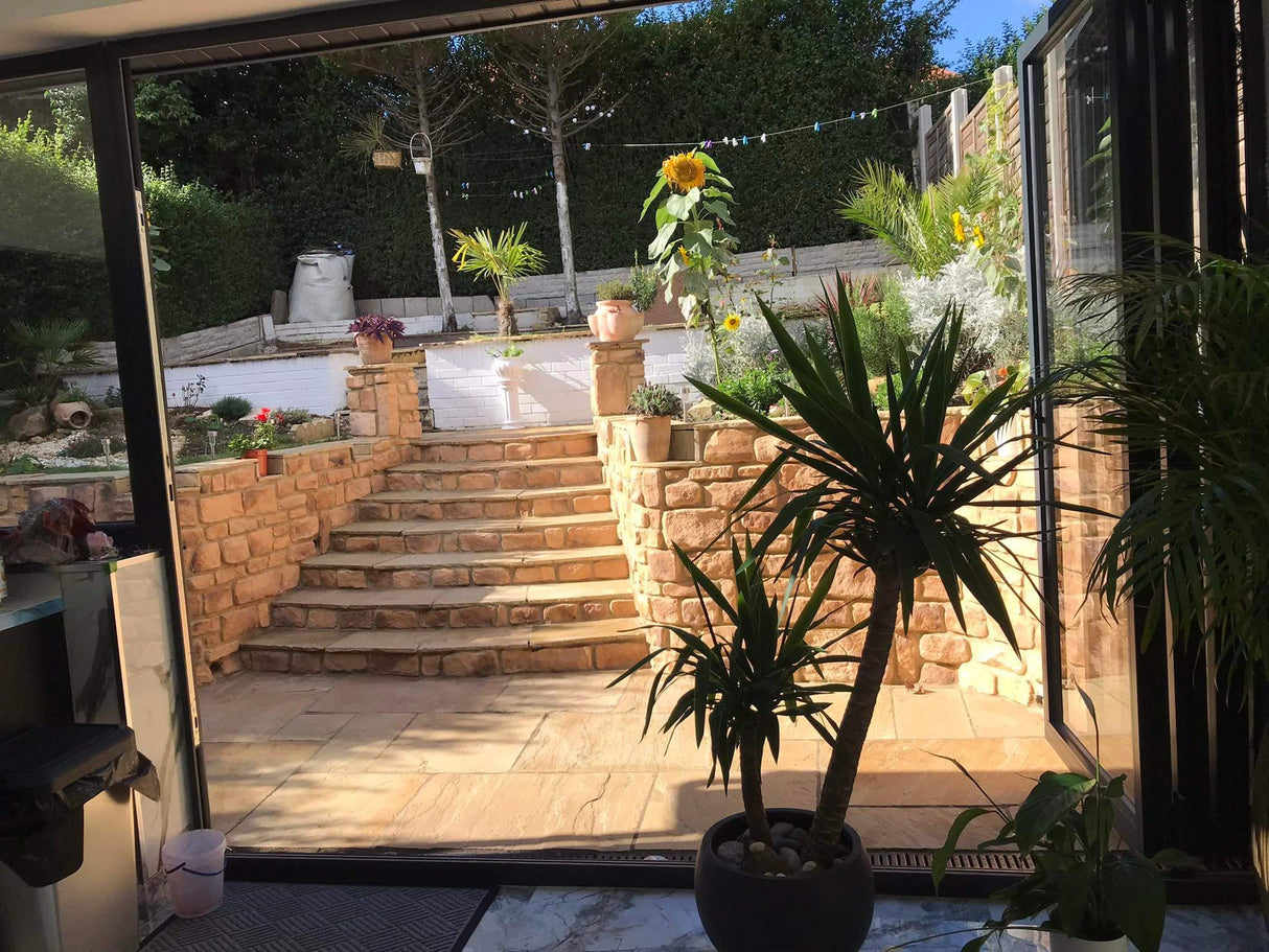 View of a sunny garden with stone steps, plants, and a patio seen through a large glass door.